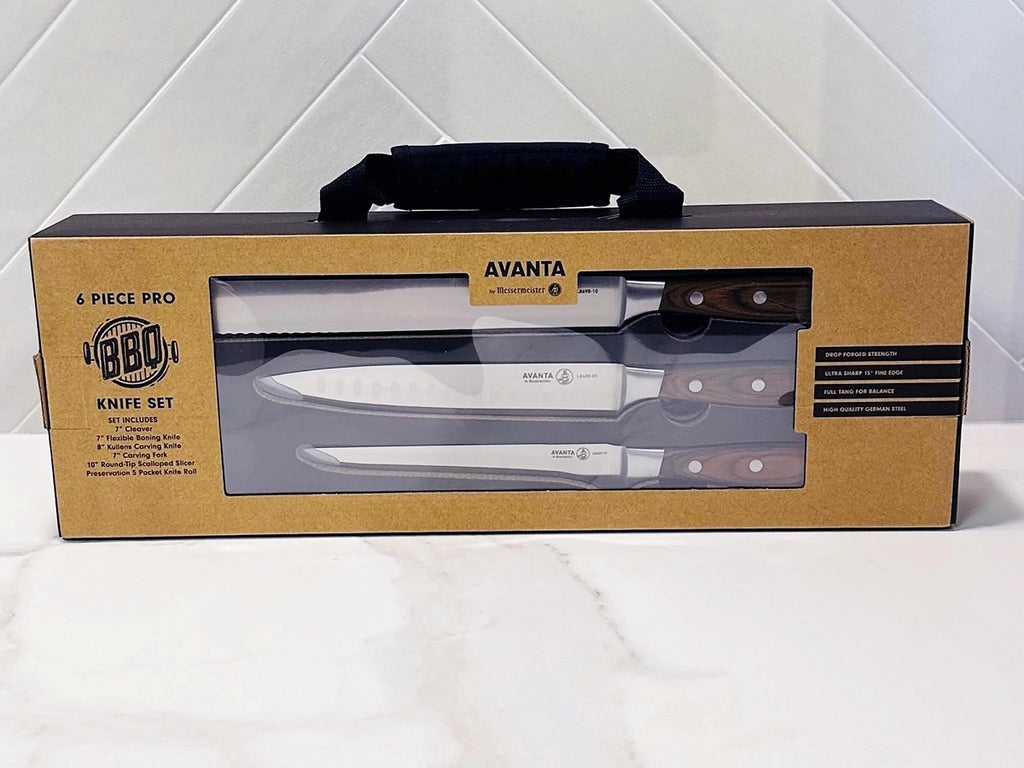 Messermeister Avanta BBQ Knivsett, et 6-delt sett med trehåndtak, vises i eske på en hvit flate foran en flislagt vegg. Den brune emballasjen har et gjennomsiktig vindu som viser Messermeisters grillkniver på innsiden.