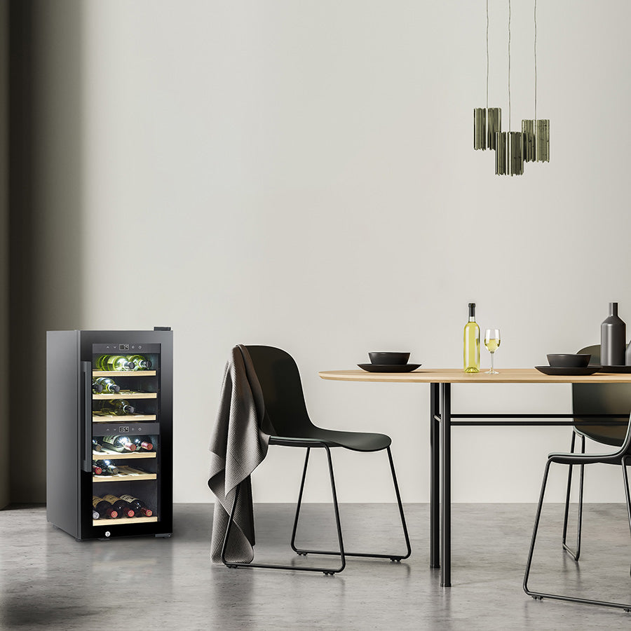 En moderne spiseplass med et elegant, svart Caso WineExclusive 24 Smart frittstående vinkjøleskap med to temperatursoner og app-styring via Wi-Fi, to stoler, et bord med glass og flasker, hengende lamper og en grå vegg i bakgrunnen.