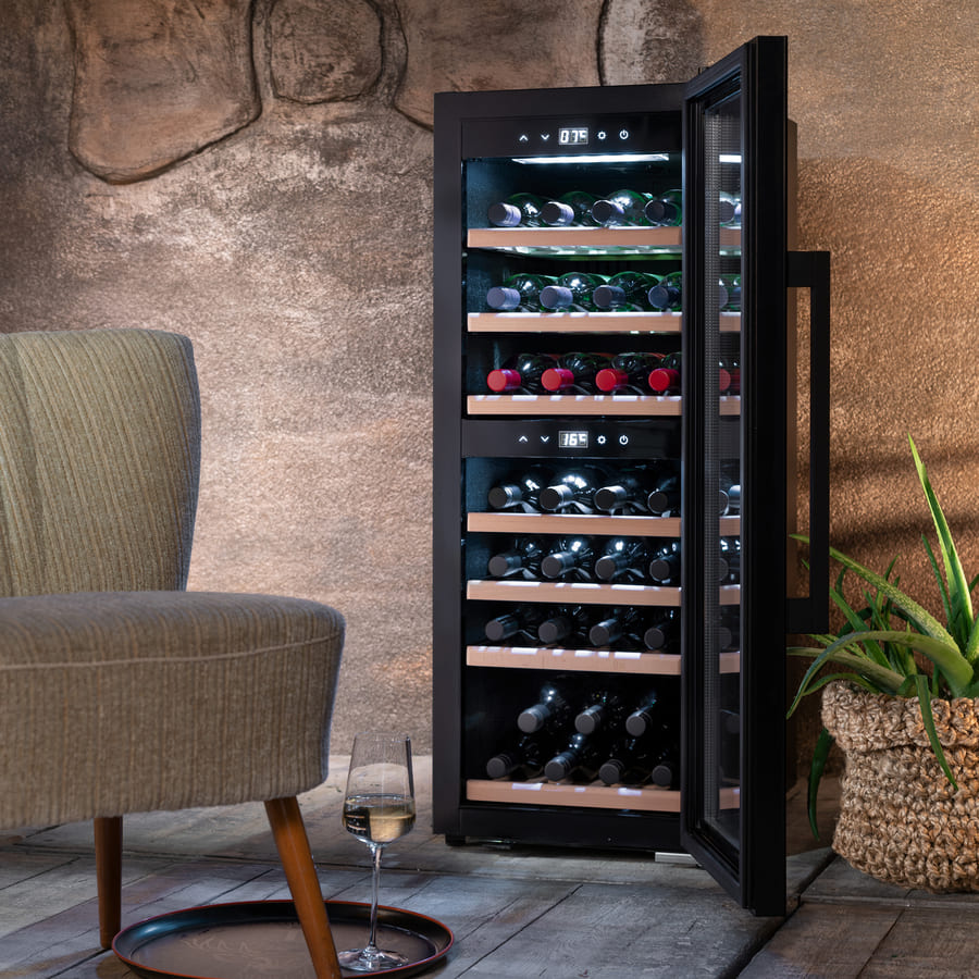 Caso WineExclusive 38 Smart Falstaff Edition, et frittstående vinkjøleskap med to temperatursoner og en glassdør, står åpent og viser frem flere flasker på trehyller. I nærheten står en stol, et brett med et vinglass og en potteplante.