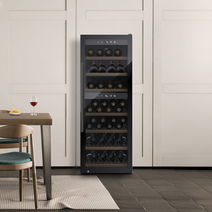 Et frittstående vinskap av modellen Caso WineExclusive 126 Smart Falstaff Edition med glassdør er fylt med flasker. Et glass rødvin står på et bord ved siden av en stol i et moderne kjøkken med lyse skap, og vinskapet har to temperatursoner for optimal lagring.