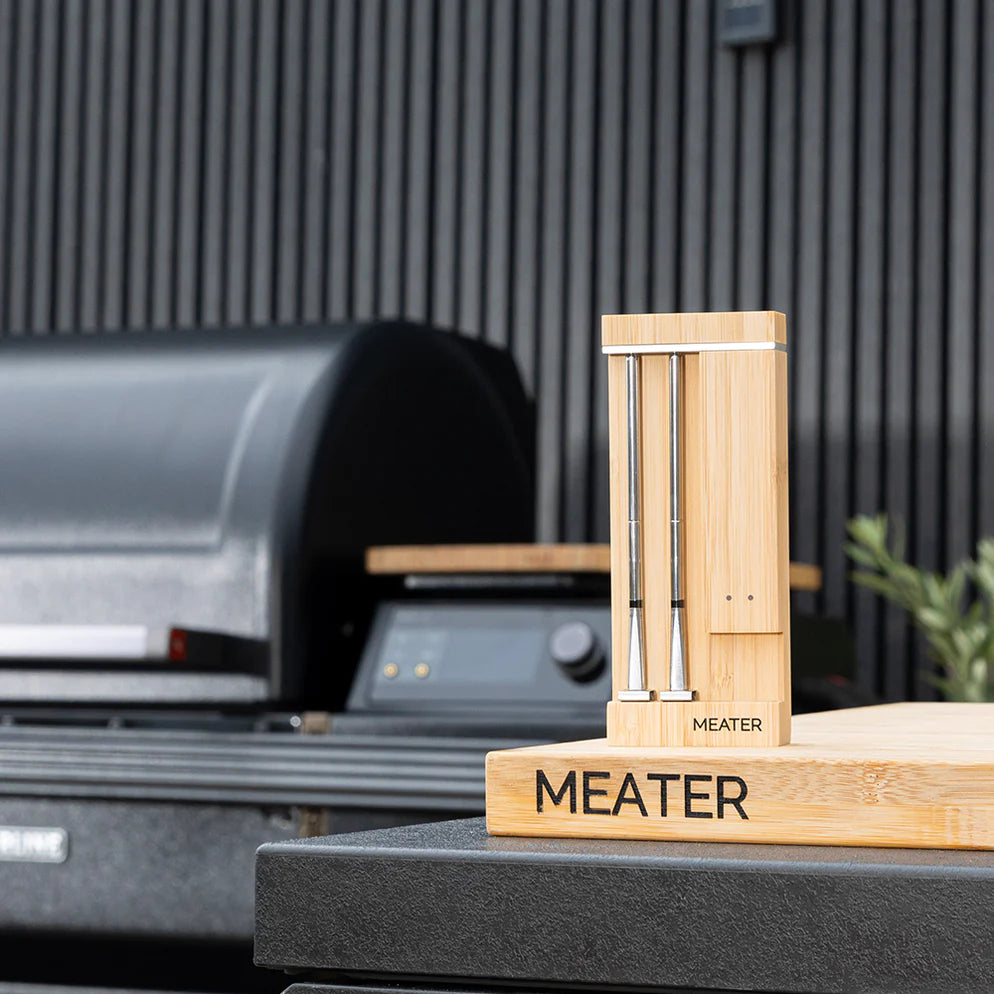 To Traeger Meater Pro Duo WiFi-steketermometre står i en MEATER-merket ladeblokk av tre på en utekjøkkenbenk i nærheten av en svart grill.