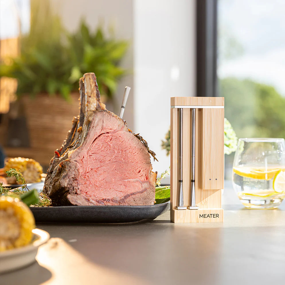 På bordet står en stekt ribbe med et Traeger Meater Pro Duo - WiFi-steketermometer på plass, ved siden av understellet og et glass vann med sitronskiver i bakgrunnen.