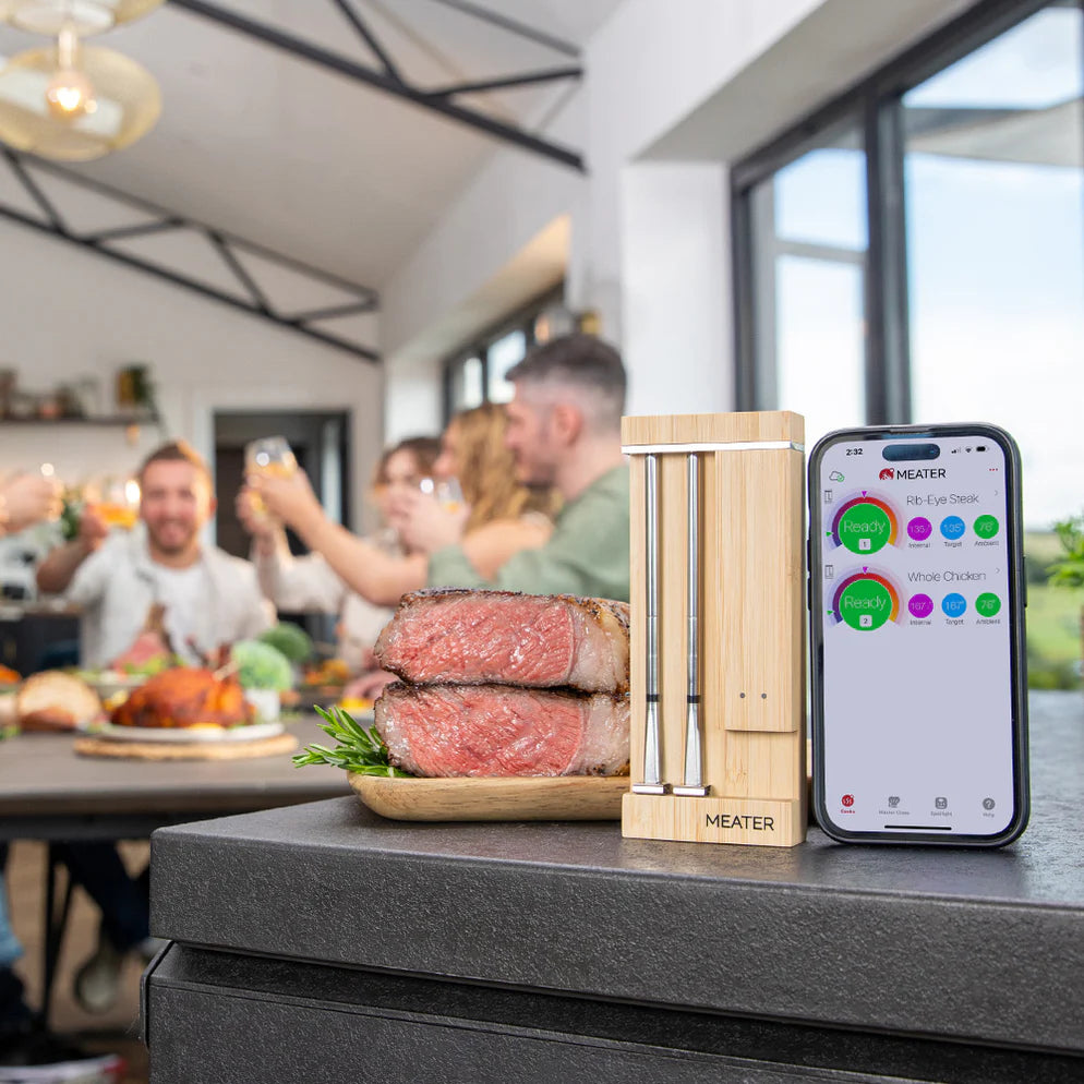En oppskåret biff ligger på en treplate ved siden av Traeger Meater Pro Duo - et WiFi-steketermometer og en smarttelefon som viser steketemperaturen, mens gjestene nyter måltidet i bakgrunnen.