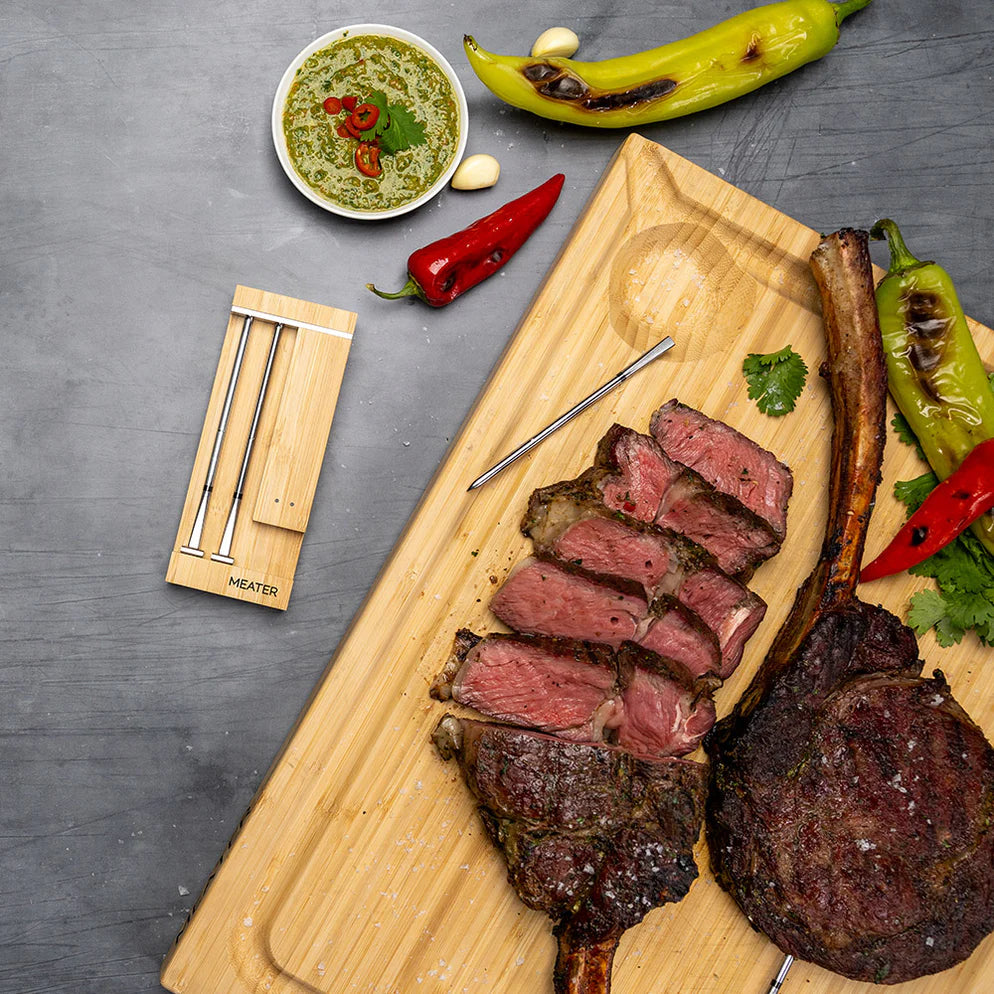 Skiver av medium stekt tomahawk-biff med grillet paprika, urtesaus og krydder på et trebrett ligger ved siden av Traeger Meater Pro Duo - WiFi-steketermometer på en grå overflate.