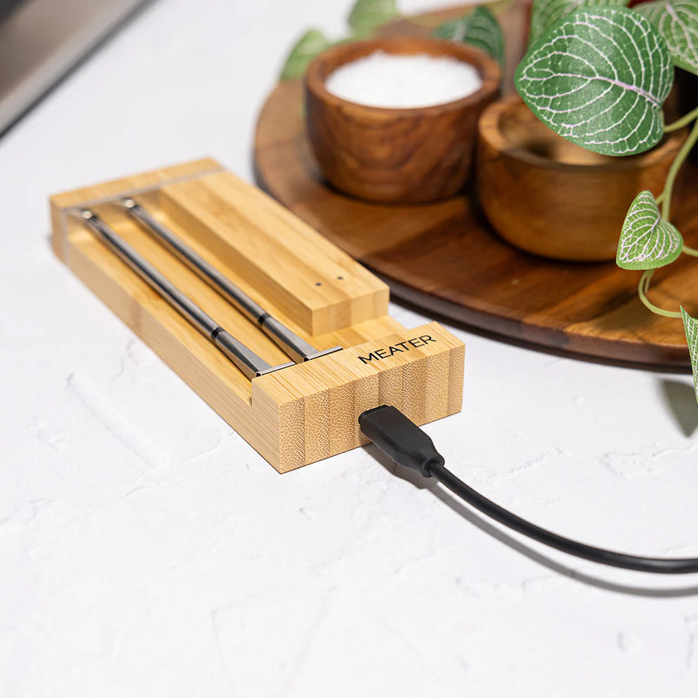 Et Traeger Meater Pro Duo WiFi-steketermometersett hviler på en ladestasjon av bambus med en svart kabel, utstilt på en hvit benkeplate ved siden av en plante og treskåler.