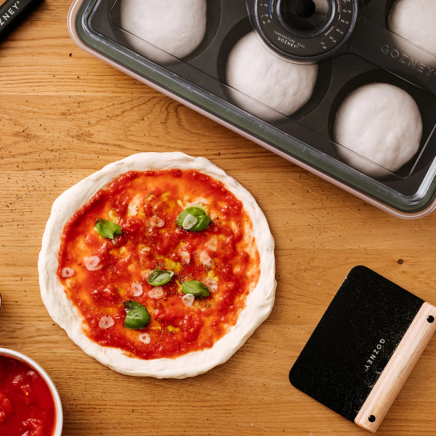 Ukokt pizzadeig med tomatsaus og basilikum på en treflate, omgitt av deigkuler, en bolle med saus, en deigskrape og Gozney Dough Tray - Hevekasse fra Gozney for optimal heving og fermentering.