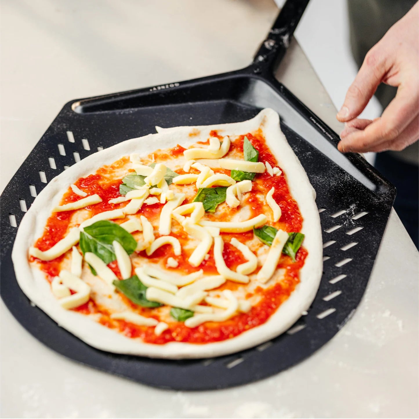 En rå pizza med tomatsaus, ost og basilikum ligger på en Gozney Balance Pizzaspade 14" Large fra Gozney, og det perforerte aluminiumsbladet er klart til baking mens noen griper om det ergonomiske håndtaket.