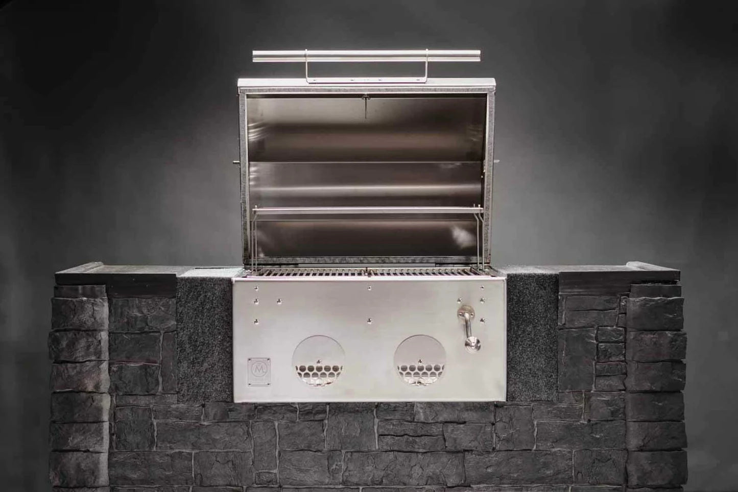B24 Kullgrill av rustfritt stål fra M Grills er innebygd i en mørk steinramme med åpent lokk, synlige grillrister, justerbar kullrist og kontrollknapper.