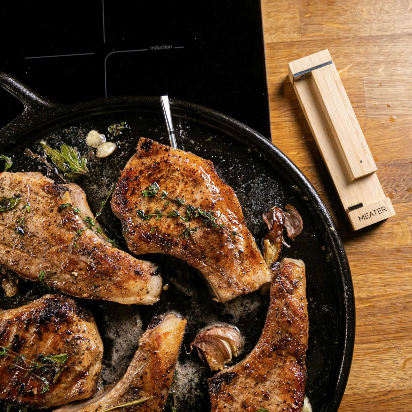 Tre stekte svinekoteletter med urter og hvitløk i en støpejernspanne, den ene med kjøtttermometer, på en trebenk ved siden av Traeger Meater Pro - Bluetooth steketermometer.