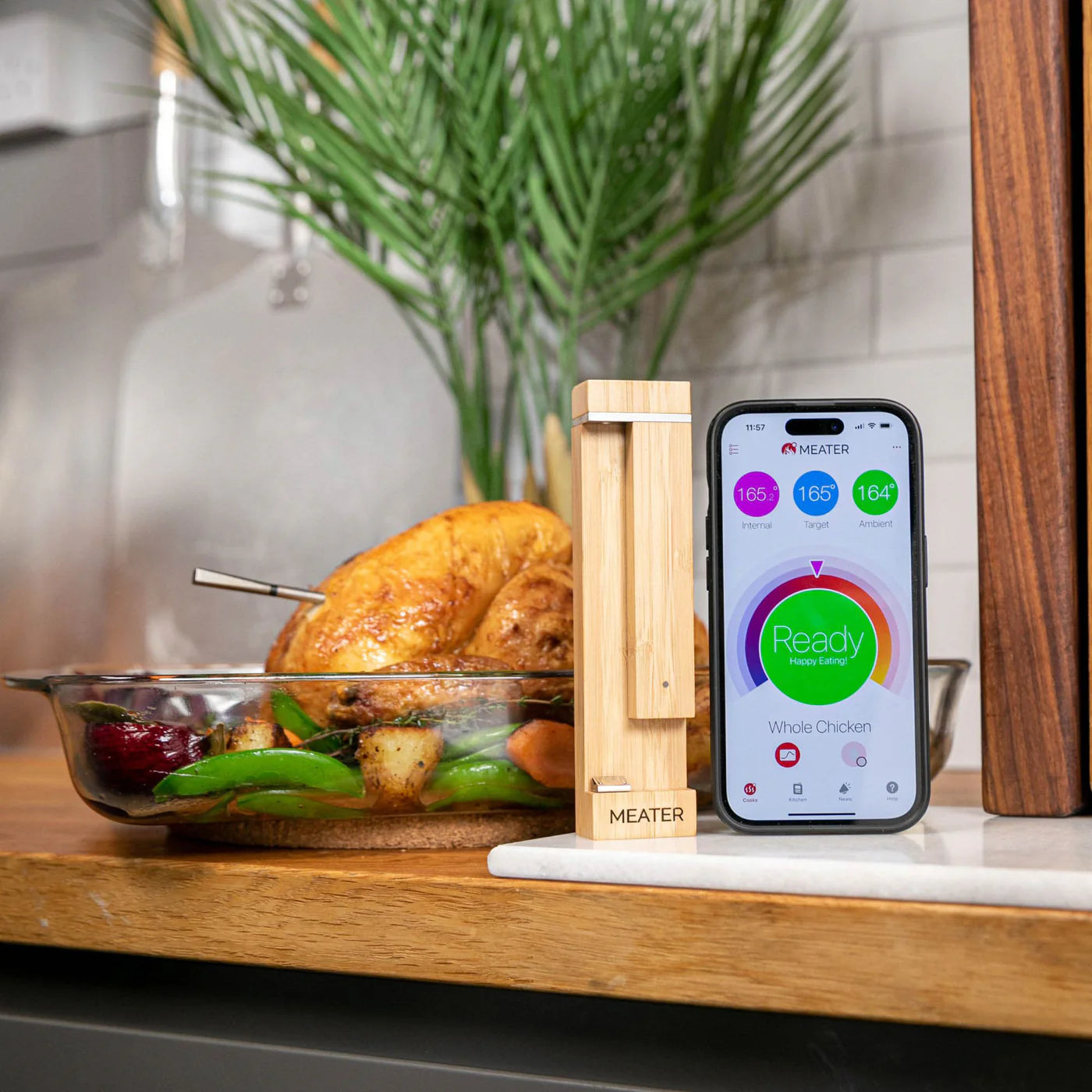 En helstekt kylling med grønnsaker i et glassfat ligger ved siden av en smarttelefon som viser en app med kjøtttermometer og Traeger Meater Pro - Bluetooth-dockingstasjon for steketermometer.