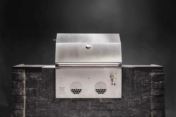 B24 Stainless Steel Built In Charcoal Grill fra M Grills er en elegant kullgrill av rustfritt stål med justerbar kullrist og temperaturmåler, flott innebygd i robust steinmur mot en mørk bakgrunn.