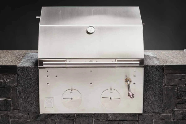 B24 Stainless Steel Built In Charcoal Grill fra M Grills - rustfritt stål kullgrill med lukket lokk og temperaturmåler, innebygd i steinbenk mot mørk bakgrunn.