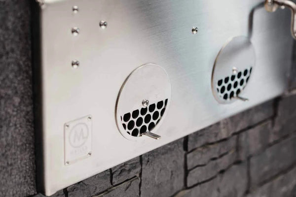 Nærbilde av M Grills B24 Built In Charcoal Grill i rustfritt stål, med et perforert ventilasjonspanel i bikakestruktur med to sirkulære ventilasjonsåpninger, montert på en steinvegg.