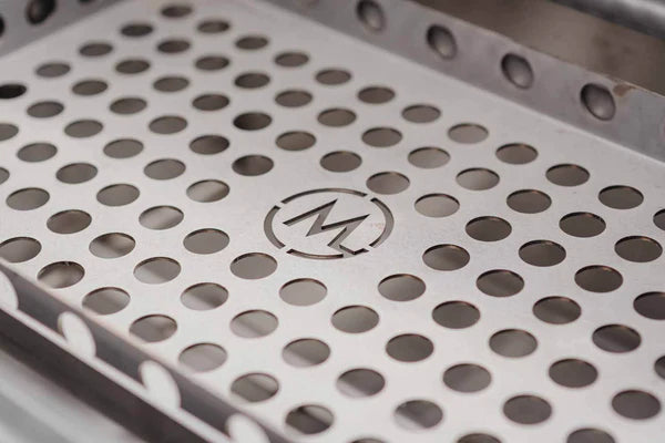 Nærbilde av et perforert metallbrett designet for B24 Stainless Steel Built In Charcoal Grill fra M Grills, med jevnt fordelte sirkulære hull og en inngravert M-logo i midten.