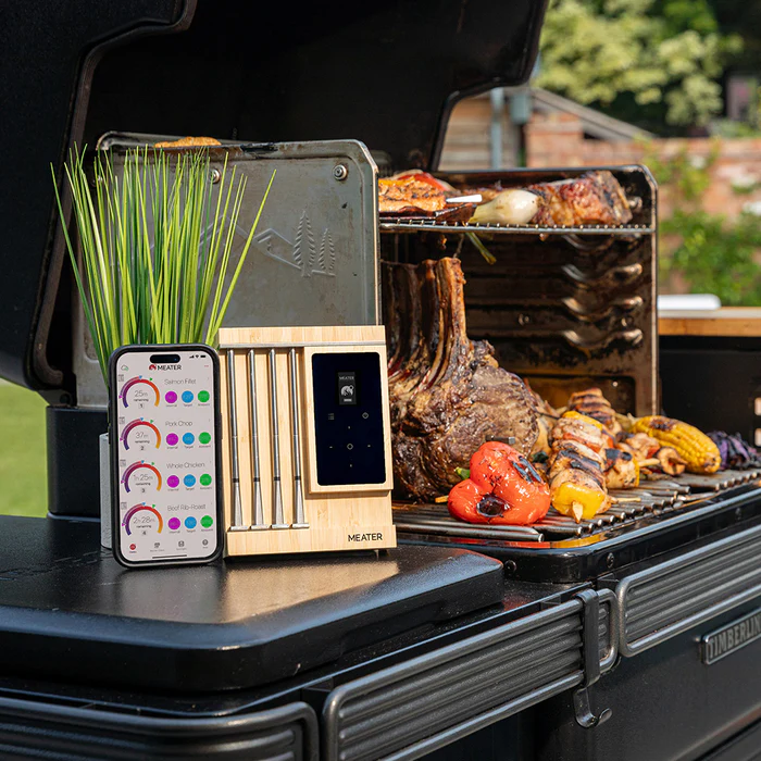 Et Traeger Meater Pro XL - WiFi-steketermometer står ved siden av en smarttelefon i nærheten av en utegrill fylt med diverse kjøtt og grønnsaker.