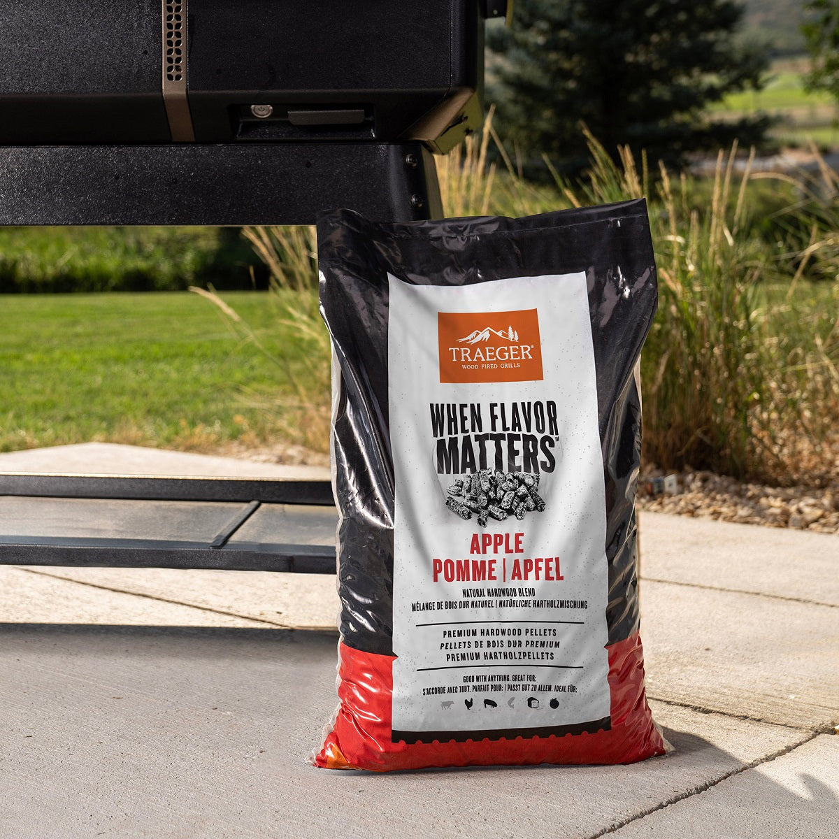 En 9kg sekk Traeger Apple Pellets fra Traeger, laget av 100% rent tre, står ved grillen på terrassen med grønt gress og trær i bakgrunnen - klar til å gi maten autentisk røyksmak.