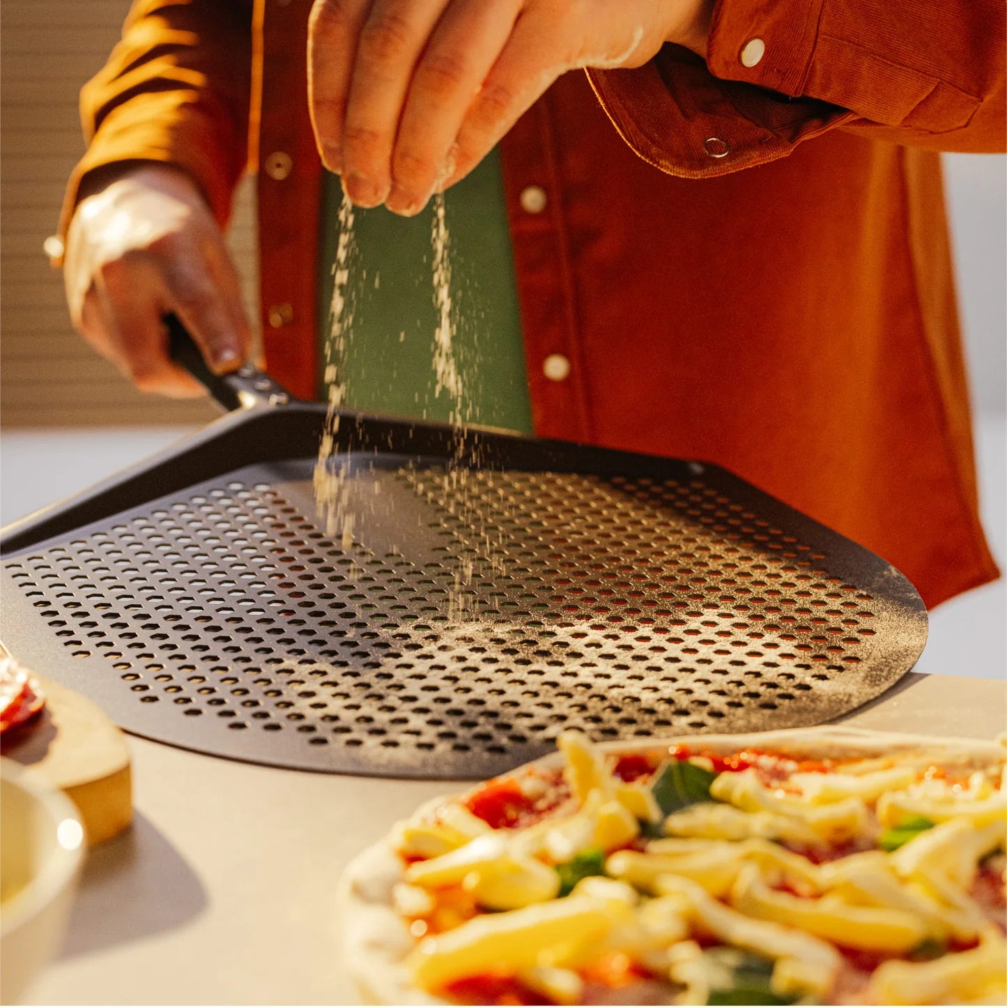 En person strør mel over en Gozney Pro Pizzaspade, 12" Medium, med en ubakt pizza toppet med ost og grønnsaker på benken foran.