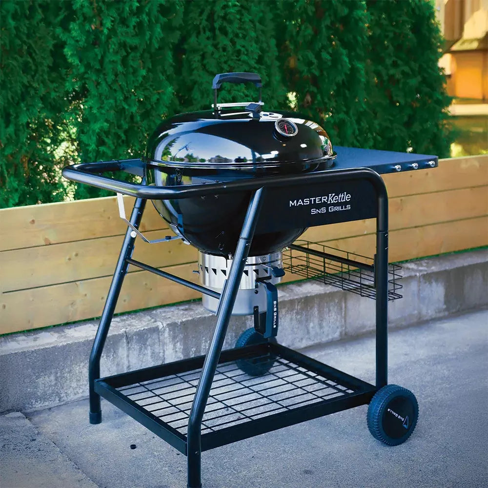 En svart SNS Master Kettle Grill 57 cm - Kulegrill med lokk, sidehylle og hjul er plassert utendørs ved et tregjerde og grønne busker - et perfekt sted å nyte allsidig kullgrilling med Slow 'N Sear-systemet.