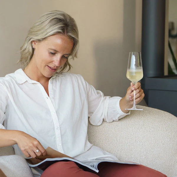 En kvinne sitter på en lys stol og leser et magasin med den ene hånden, mens hun holder et Stölzle Lausits STARlight champagneglass 290 ml fylt med musserende vin i den andre.