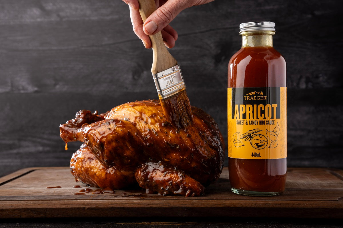 En hånd pensler BBQ-saus på stekt kylling, med en flaske Traeger Apricot BBQ Sauce 440 ml fra Traeger ved siden av - perfekt pølse til kylling og svin - klar til å nytes som den ultimate grilldipp på en treflate.