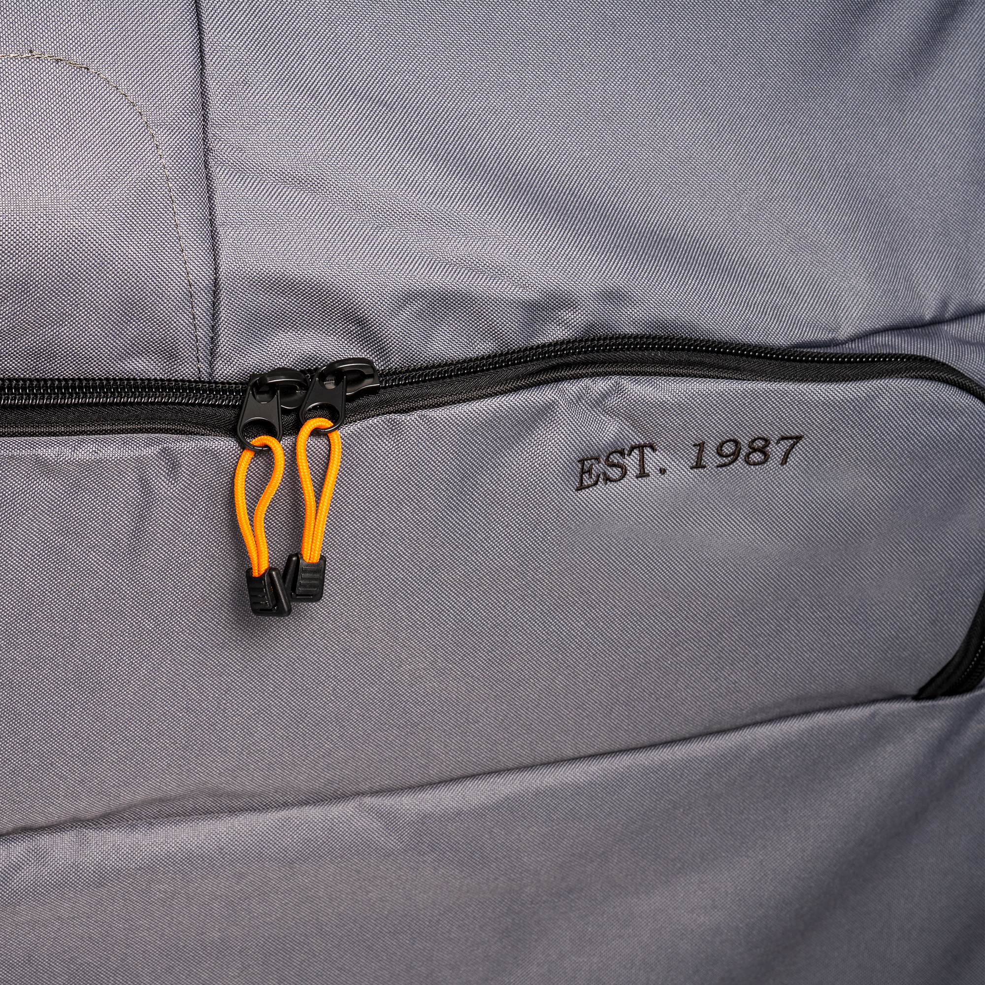 Nærbilde av en grå stoffpose med svart glidelås, oransje trekk og "EST. 1987"-broderi - ideell for oppbevaring av Traeger Full Length Grill Cover for Timberline L fra Traeger.