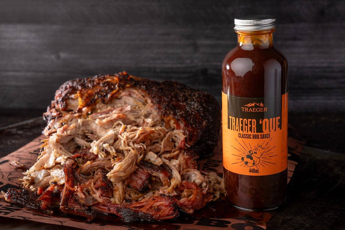 En haug med strimlet svinekjøtt ligger ved siden av en flaske Traeger Que BBQ Sauce 440 ml fra Traeger, og den røykfylte aromaen lokker på brunt papir mot en mørk trebakgrunn.