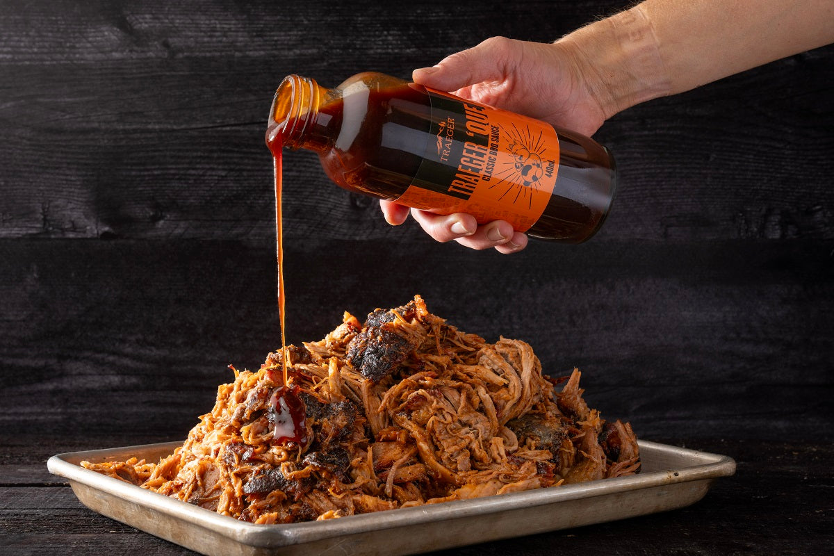 En hånd heller Traeger Que BBQ Sauce 440ml fra Traeger på et brett med strimlet, stekt kjøtt mot en mørk bakgrunn.