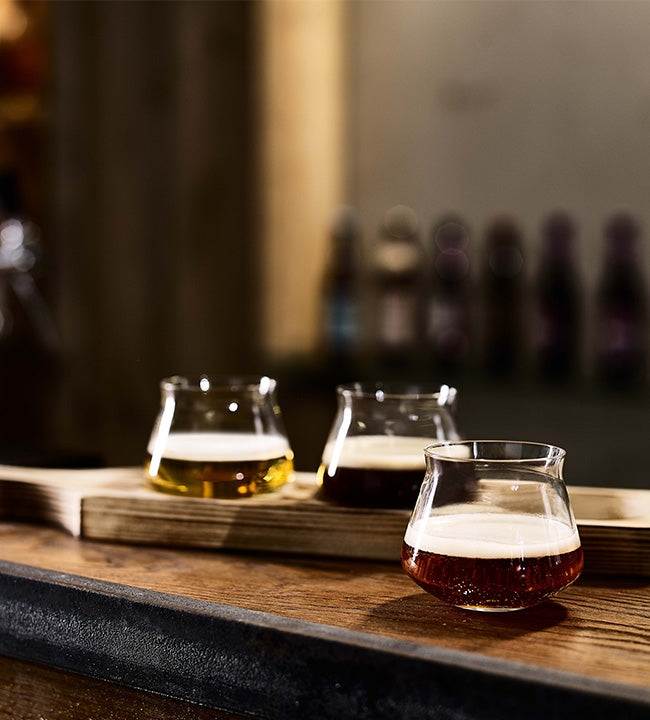 Tre små øl i Rastal Teku Taster Flight Set - smaksglass og smaksplanke er anrettet på bardisken, med en mykt uskarp bakgrunn.
