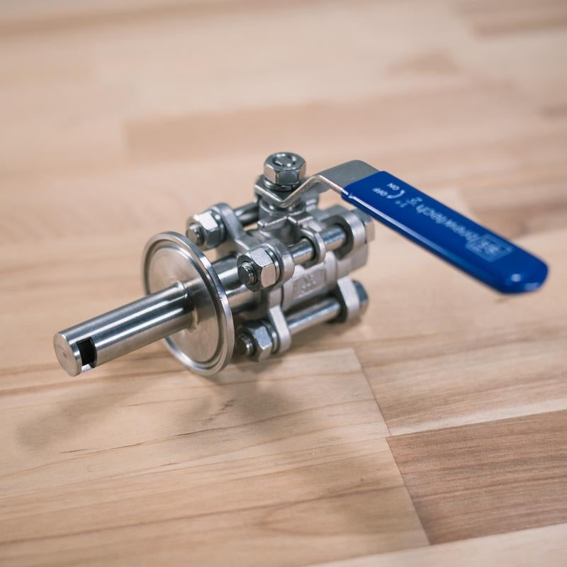 Ss Brewtech 1,5" Tri-Clamp Whirlpool Fitting i rustfritt stål med blått håndtak og tri-clamp tilkobling hviler på en overflate av tre.