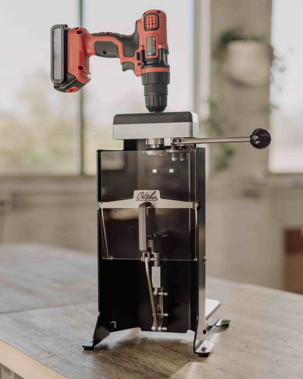 En rød batteridrevet drill er montert på toppen av BenchMK Drill-Powered Can Seamer fra Oktober Design, som har et gjennomsiktig frontpanel og er plassert i et godt opplyst rom.