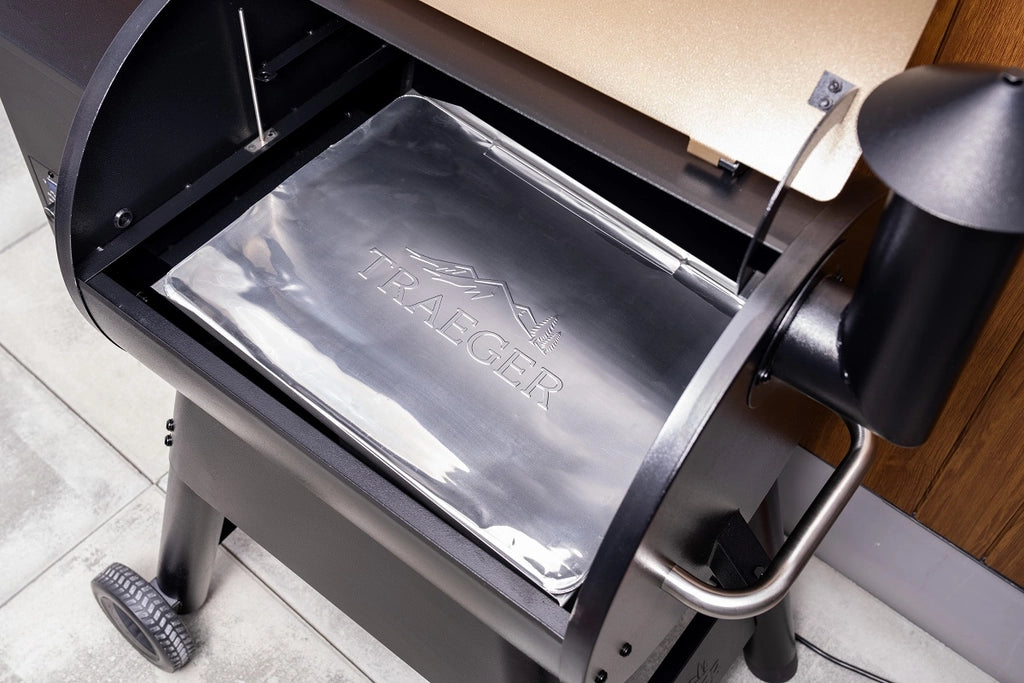 Et nærbilde av en åpen Traeger-grill viser et skinnende dryppbrett og fettoppsamler, perfekt for bruk med Traeger Drip Tray Liners størrelse A 5-pk Pro22/Pro575/IW650/Timberline 850 - folieunderlag og ditt favoritt grilltilbehør.