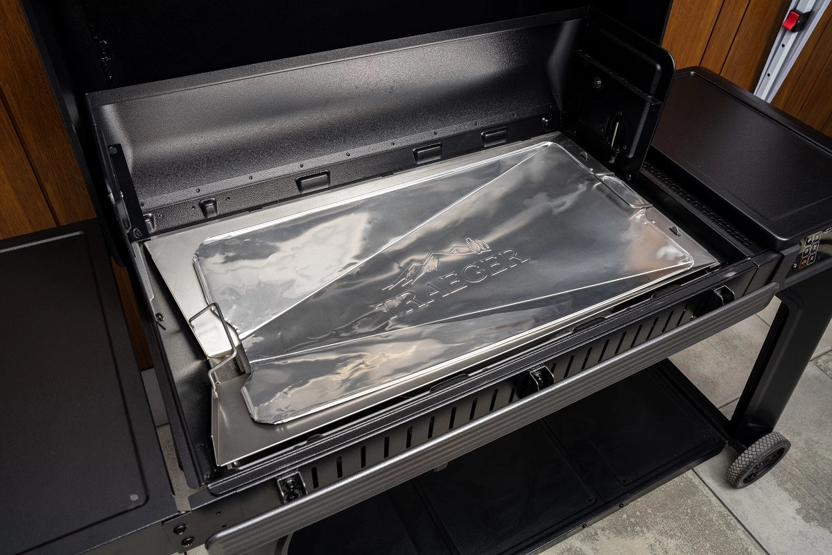 Nærbilde av en grill med et skinnende brett preget med TRAEGER, med Traeger Drip Tray Liners, størrelse C 5-Pack Woodridge Elite/Timberline 1300 - folieunderlag, som uunnværlig tilbehør for enkel rengjøring.