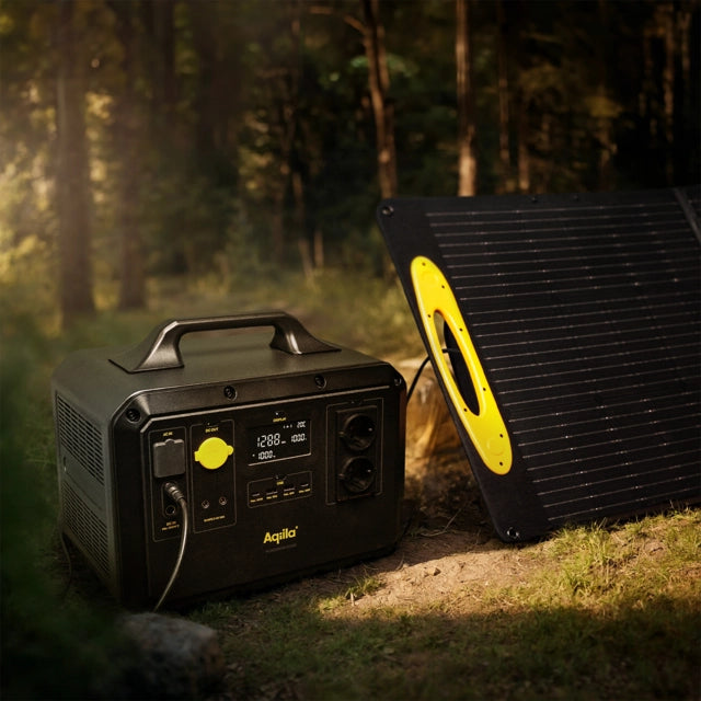 Et Aqiila Powerbird S1500 - Kraftstasjon nødbatteri fra Aqiila er koblet til et solcellepanel som er plassert på bakken i en solfylt skogsgløtt.