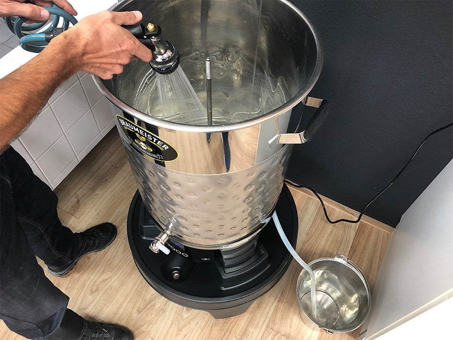 En person bruker en spraydyse til å rengjøre en stor bryggekjele i rustfritt stål, som står på en Speidel Base med hjul til Braumeister 50L, ved siden av en metallbøtte som samler opp vann.