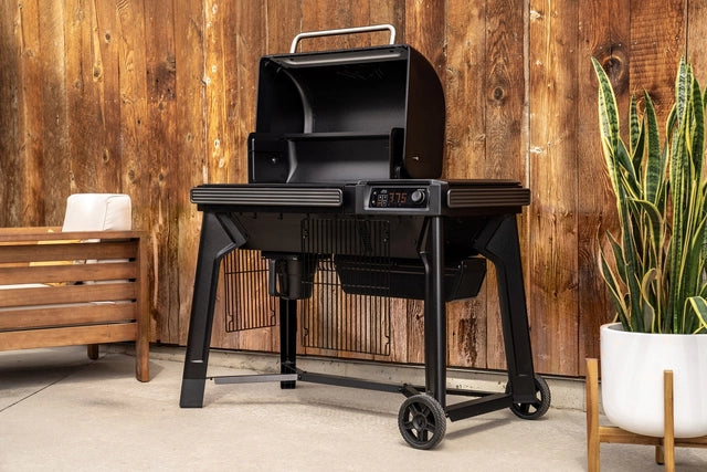 En elegant, svart Traeger Woodridge pelletsgrill med WiFIRE-teknologi og digitalt display står på terrassen ved en trebenk og potteplante, og byr på ekte vedfyrt grillsmak ved siden av en trevegg.
