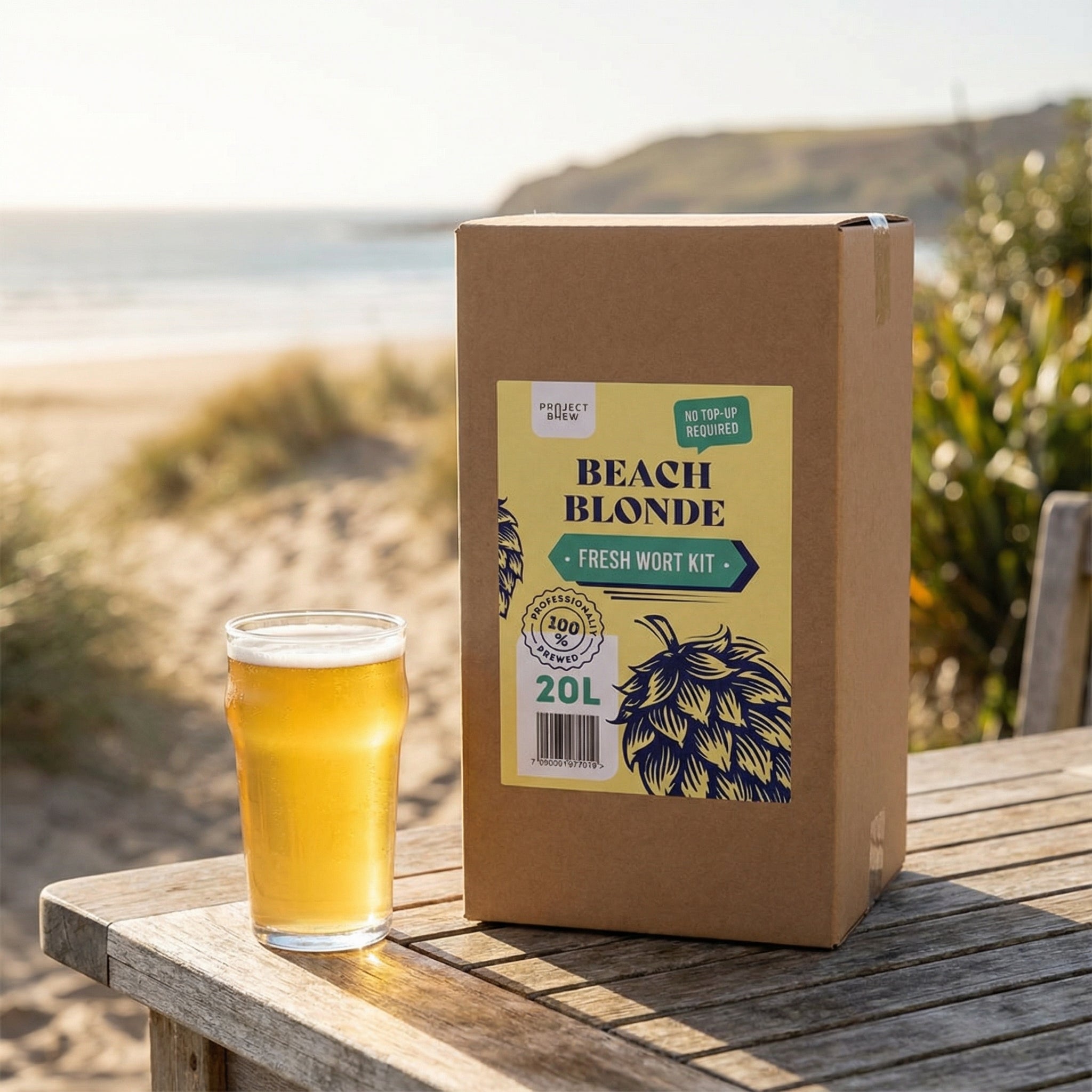 På et trebord utendørs nær stranden står en eske merket "Beach Blonde - 20L Fresh Wort Kit" fra Austmann og en halvliter amerikansk Blonde Ale.