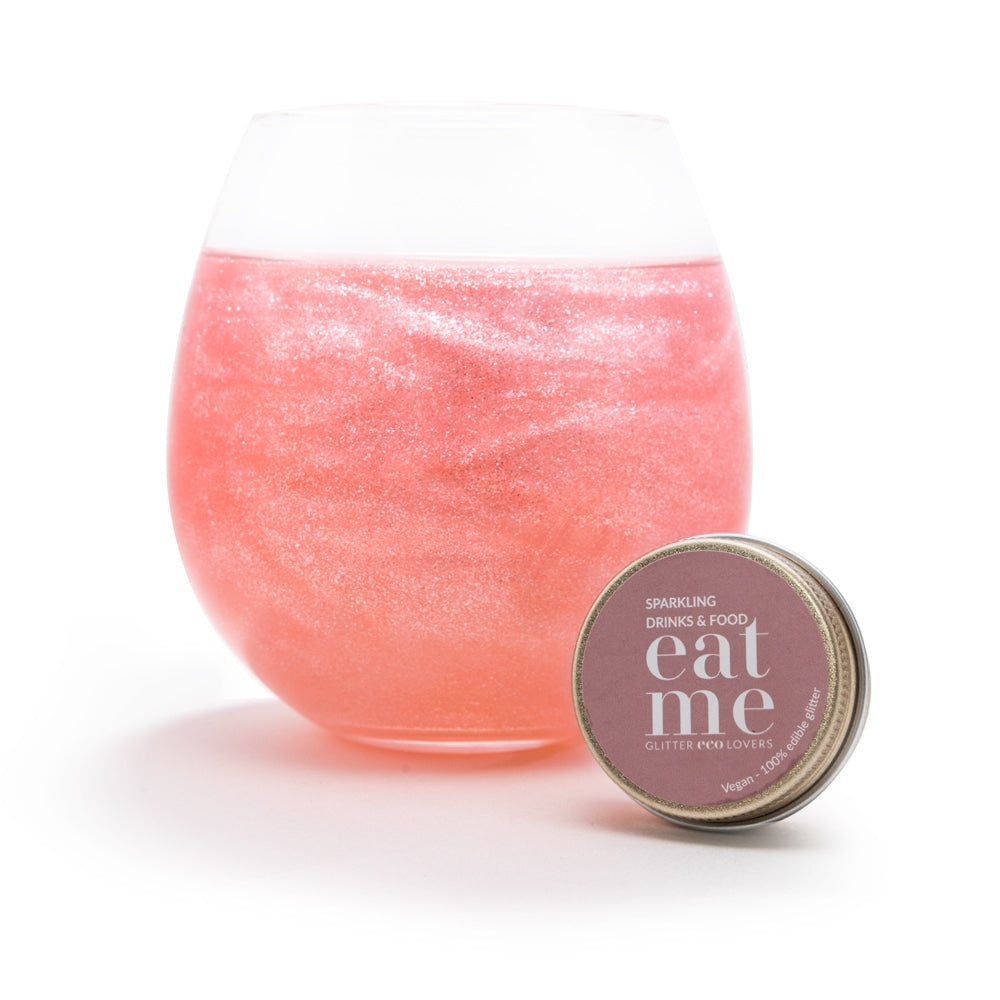 Et rundt glass med rosa, glitrende drikke glitrer med Eco Lovers Eat Me Glitter Rose 20 ml, ved siden av en liten beholder merket "eat me sparkling drinks & food glitter".