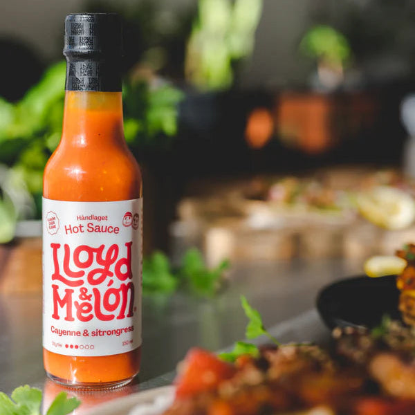 En flaske Cayenne & Sitrongress Hot Sauce | Lloyd & Melón, laget med naturlig fermentering, står på et bord ved siden av tallerkener med mat, med frodig grønt i bakgrunnen.