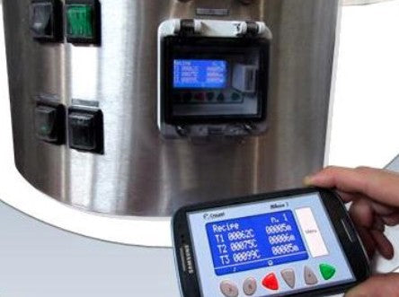 En person bruker et Camurri Bluetooth-grensesnitt fra Camurri i nærheten av en maskin i rustfritt stål som er utstyrt med brytere, et digitalt display og støtte for enkel installering for brukervennlighet.