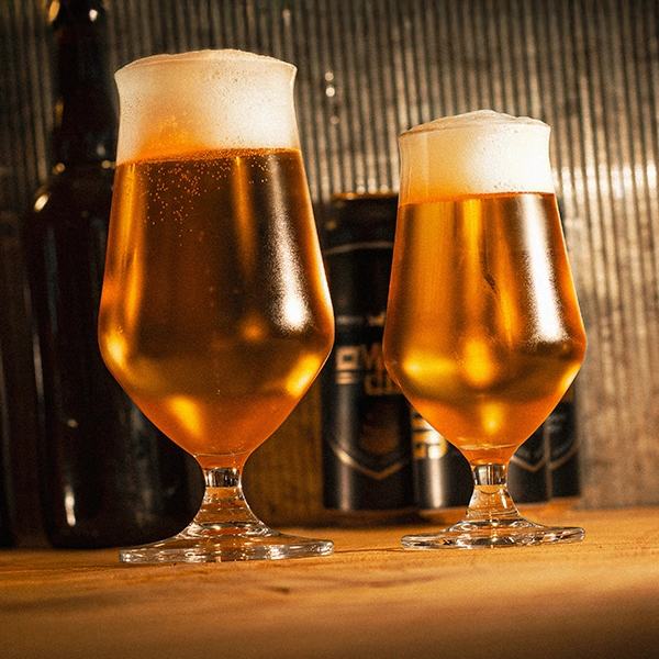To Rastal Craft Master Taste 37cl ølglass fra Rastal, fylt med ravfarget øl og tykke skumhoder, står på en treflate mens uskarpe flasker og bokser dukker opp i bakgrunnen.