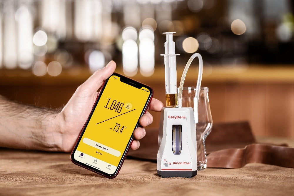 En person holder en smarttelefon som viser målingene fra EasyDens Digitalt Hydrometer fra Anton Paar, ved siden av en digital alkoholmåler og et glass øl og vin på et bord i en bar eller restaurant.