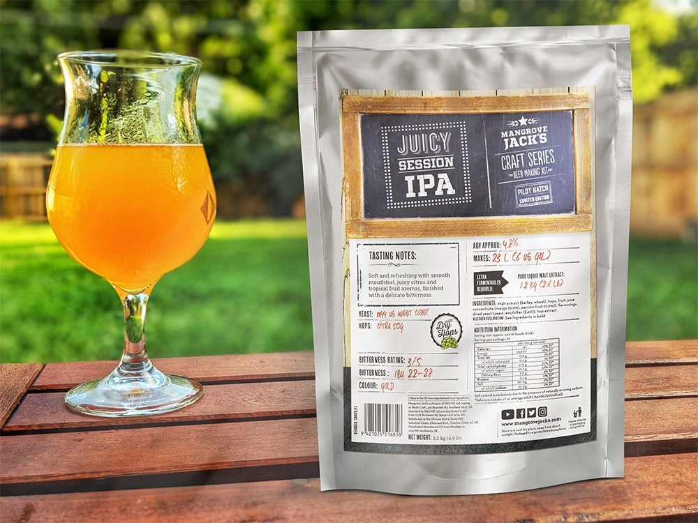 Et glass oransje øl står på et trebord ved siden av en Mangrove Jacks Craft Series Juicy Session IPA ekstraktsett - 2,2 kg, som har detaljert produktinformasjon og maltekstrakt av høy kvalitet på emballasjen.