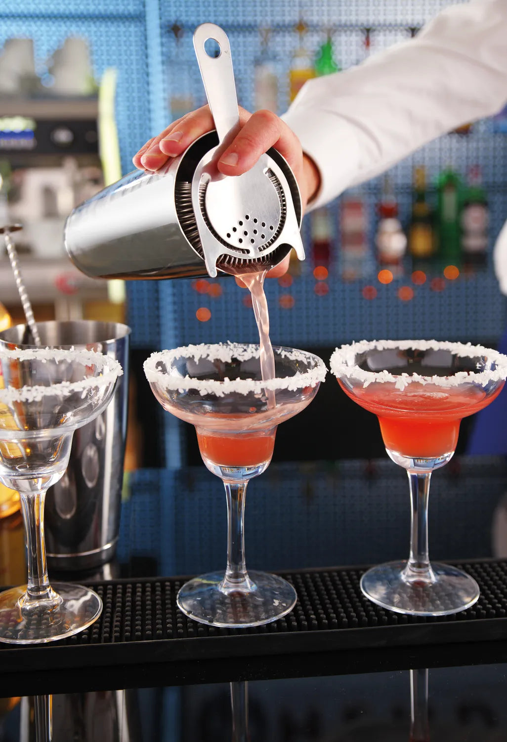 En bartender skjenker en rosa cocktail gjennom Hendi Hawthorne Strainer i margaritaglass med saltet kant, mens uskarpe barflasker fyller bakgrunnen.