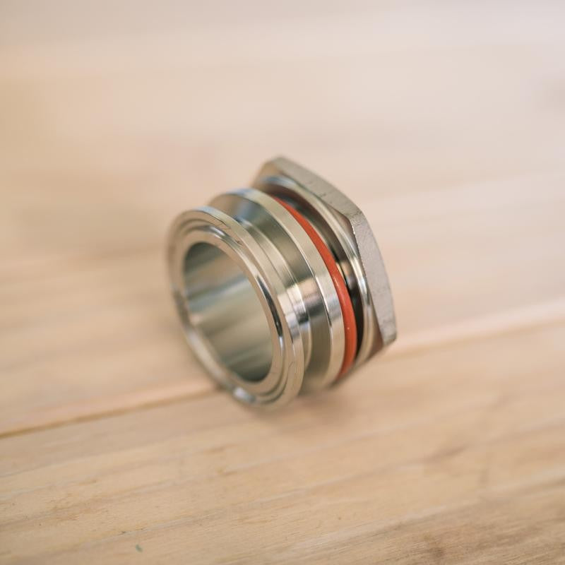 Nærbilde av Brewshop 1,5" Tri-Clamp sveisefri gjennomføring i 304 rustfritt stål med oransje pakning, utstilt på en lys treflate.