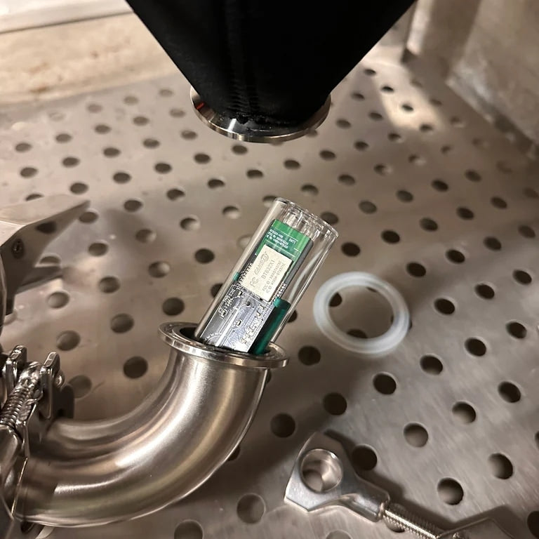 A Tilt Pro Mini Hydrometer & Termometer circuit board is enclosed in a transparent tube, inserted into a stainless steel pipe fitting on perforated metal, with nearby tools and a white circular ring visible.