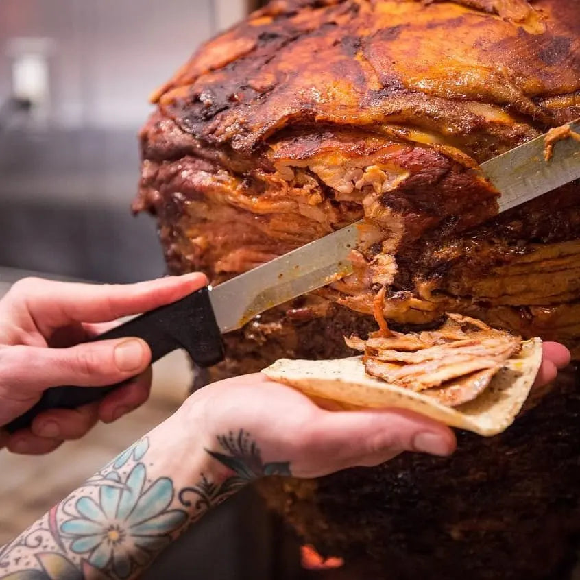 En person skjærer kjøtt fra et vertikalt spyd og legger det på en tortilla, sannsynligvis for å lage en taco. Scenen viser nærbilder av kjøttet, kniven og den tatoverte armen, mens den appetittvekkende duften av meksikansk krydderblanding og El Yucateco Achiote Flytende 300 ml lover autentiske smaker i hver eneste bit.