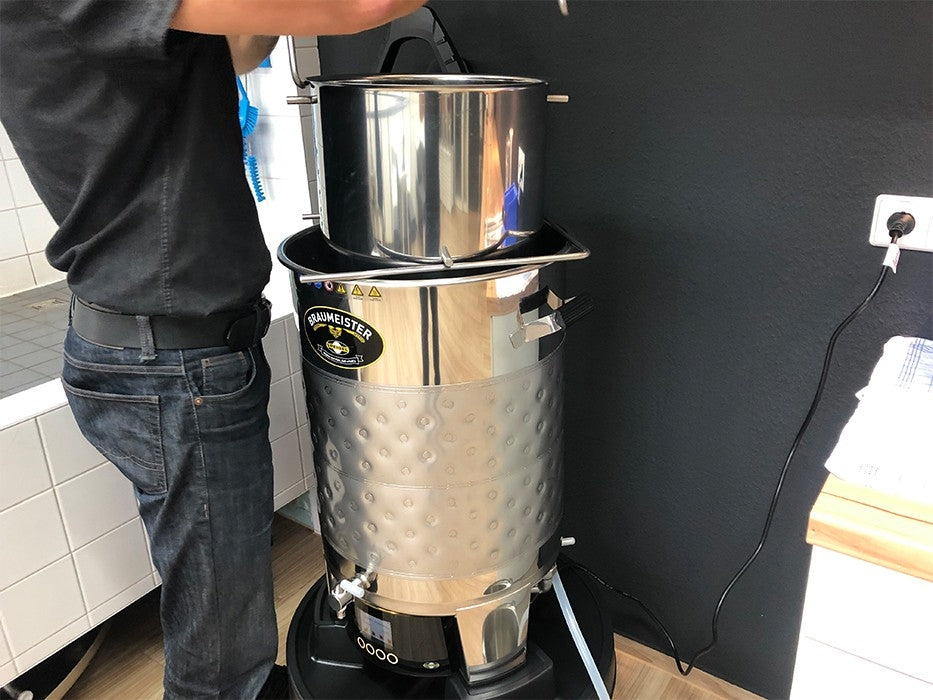 En person betjener en Speidel Base med hjul til Speidel Braumeister 50L, laget av rustfritt stål med hjul og bremser, i et flislagt rom; bryggeutstyr og et strømuttak er også synlig.