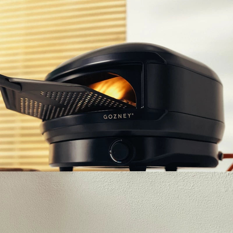 Gozney Arc Lite, Sort - Pizzaovn, har et kompakt ovn-design med flammer som gløder innvendig og en pizzaspade delvist inneni.