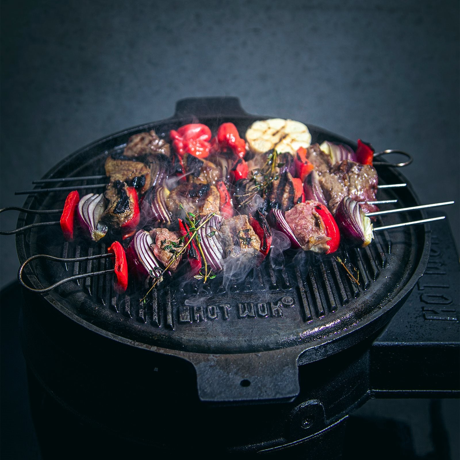 Metallspyd med kjøtt, rød paprika og løk freser over en HotWok Grillpanne 32,5 cm by HotWok gassbrennere, røyken stiger opp og en halvert hvitløkspære ligger ved siden av.