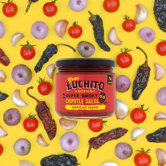 Et 330 ml glass med Gran Luchito Chipotle Salsa er omgitt av cherrytomater, halve rødløk, hvitløksfedd og tørket chili på en livlig gul bakgrunn.